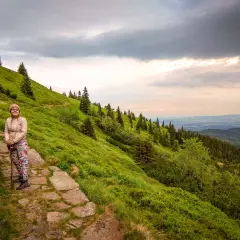 Karpacz-Śnieżka czas wejścia. Kobieta z kijkami na kamiennym szlaku, zielone zbocze górskie, chmury na niebie.