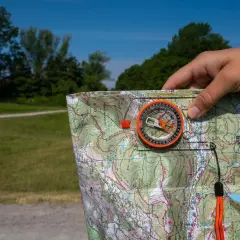 Dłoń trzyma mapę i kompas, wskazując kierunki północ południe wschód zachód. W tle zielony las i błękitne niebo.