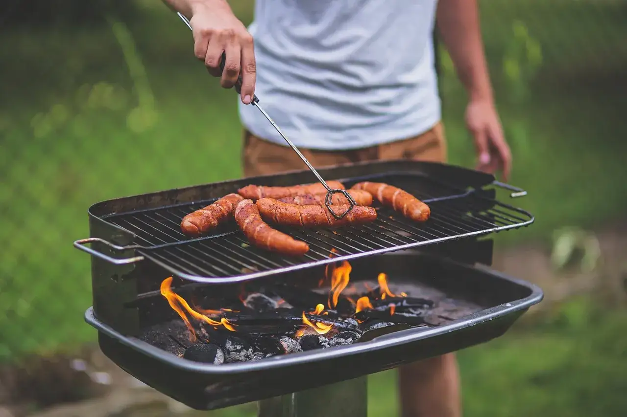 Osoba obraca kiełbaski na grillu, pokazując, jak rozpalic grilla. Płomienie żarzą się pod rusztem.