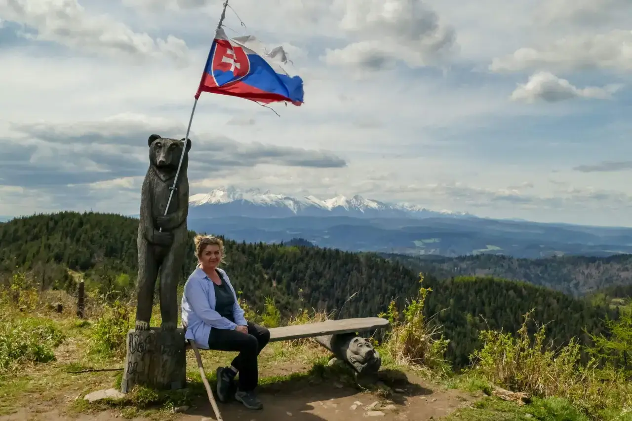 Kobieta siedzi na ławce pod słowacką flagą, obok rzeźby niedźwiedzia. Widok na g&oacute;ry z veterny vrch.
