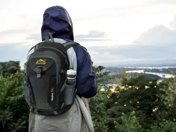 Osoba w kapturze z plecakiem gotowa na trekking górski, z widokiem na dolinę i rzekę.