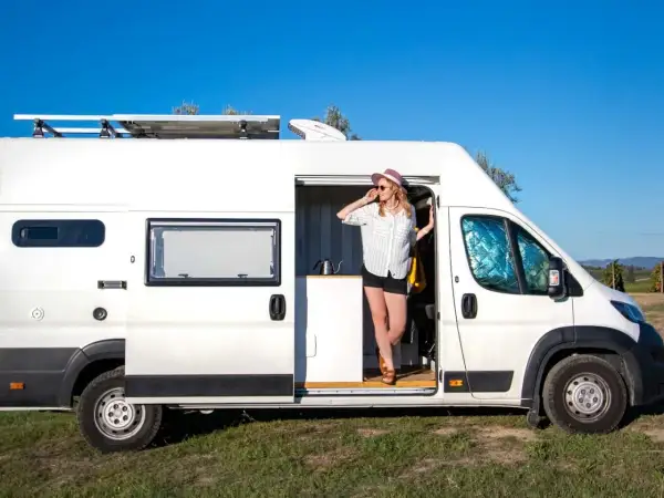 Kobieta w kapeluszu wychodzi z białego busa kampera, ciesząc się słonecznym dniem na tle błękitnego nieba.