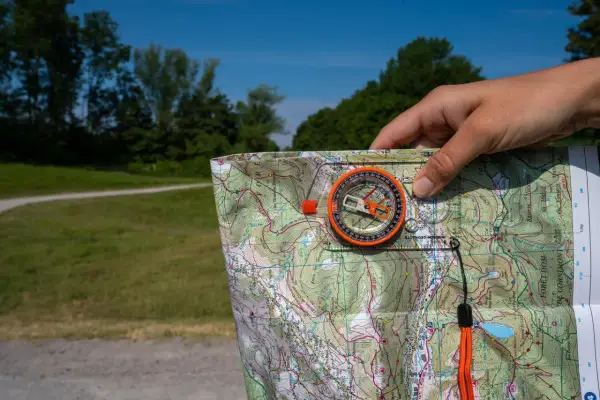 Dłoń trzyma mapę i kompas, wskazując kierunki północ południe wschód zachód. W tle zielony las i błękitne niebo.
