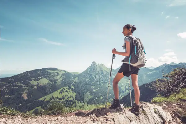 Młoda kobieta z plecakiem i kijkami trekkingowymi podziwia panoramę gór. Idealny widok dla tych, którzy zastanawiają się, jak zacząć chodzić po górach.