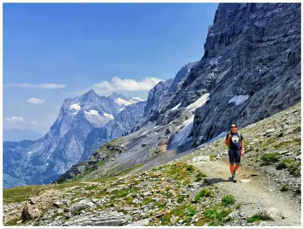 Turystka wędruje ścieżką u podnóża potężnego Eiger szczytu. W tle majestatyczne, ośnieżone góry i błękitne niebo.